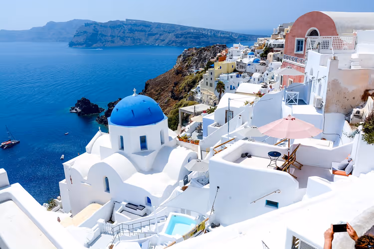 Kirche mit blauer Kuppel und weiß getünchte Terrassen auf einer Klippe über der tiefblauen Caldera von Santorin