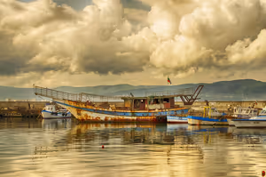 Rostiges altes Holzschiff und kleine Fischerboote in einem ruhigen Hafen unter schweren Wolken an der bulgarischen Küste
