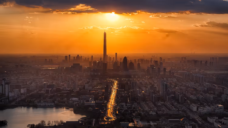 Orangefarbener Sonnenuntergang über dunstiger Stadtsilhouette mit einem hohen Turm, vorn eine beleuchtete Allee und ein Fluss