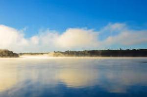 Morgennebel zieht über ruhiges Wasser, bewaldetes Ufer und Wolken spiegeln sich unter blauem Himmel