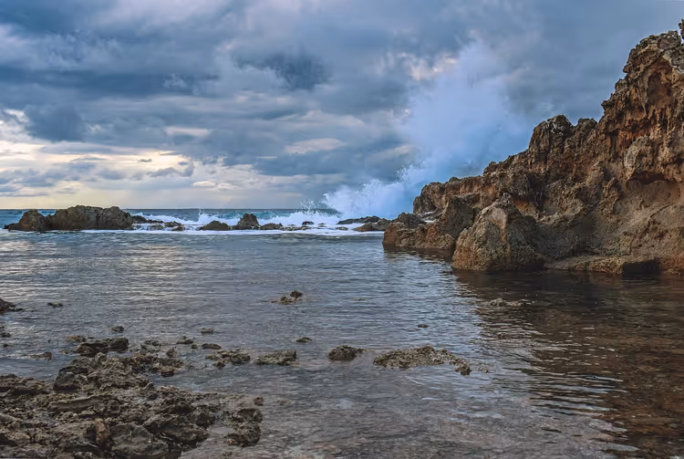 Wellen brechen sich an schroffen Küstenfelsen unter schweren Sturmwolken, im Vordergrund ruhiges, flaches Wasser