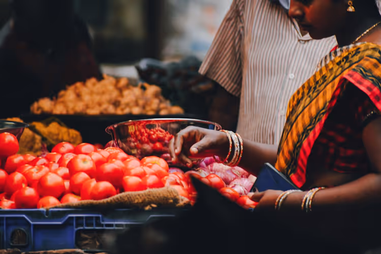 Frau im Sari mit goldenen Armreifen sucht an einem Gemüsestand auf einem Markt in Indien Tomaten und Zwiebeln aus