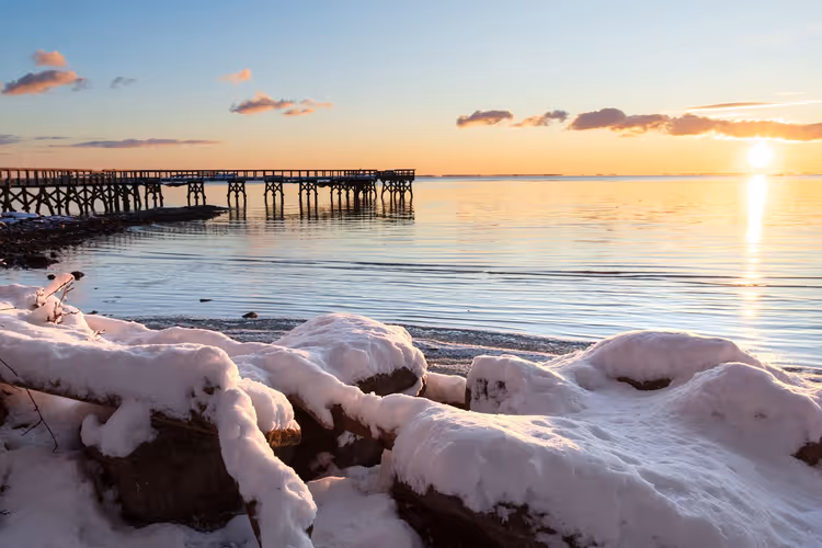 Schneebedeckte Felsen an einem ruhigen Ufer mit einem Holzsteg, der bei Sonnenuntergang ins Meer hinausragt