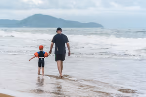 Mann und kleines Kind mit orangen Schwimmflügeln gehen Hand in Hand in die Brandung am Strand von Phuket, dahinter grüne Landzunge