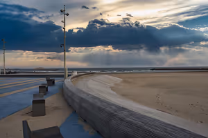Geschwungene Strandpromenade mit Bänken an einem leeren Sandstrand unter dunklen Gewitterwolken und Sonnenstrahlen