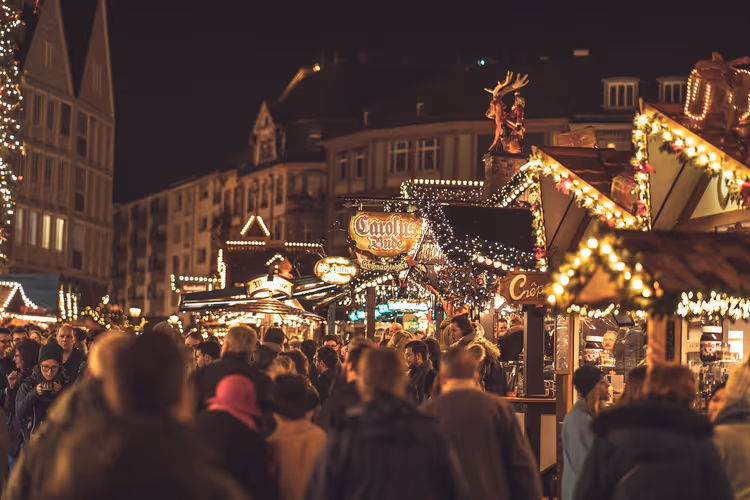 Menschenmenge zwischen beleuchteten Holzbuden auf einem Weihnachtsmarkt bei Nacht, dahinter alte Bürgerhäuser