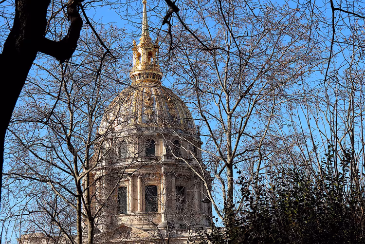 Vergoldete Kuppel und Turmspitze des Invalidendoms in Paris, durch kahle Winterzweige vor blauem Himmel gesehen