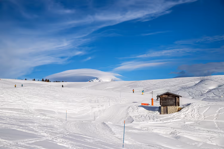 Brede besneeuwde skipiste met een kleine houten hut en een paar skiërs in de verte onder een diepblauwe lucht met sluierwolken