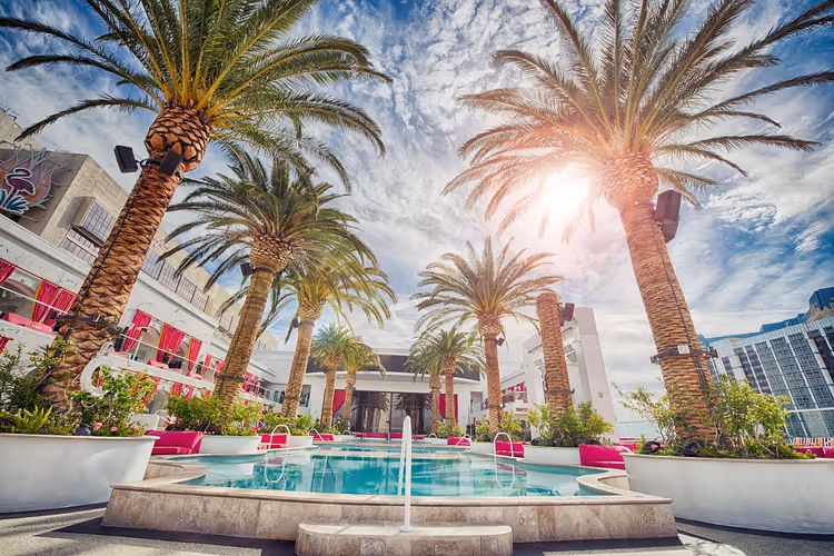 Blick von unten auf hohe Dattelpalmen rund um einen Hotelpool mit rosa Liegen und weißen Gebäuden im hellen Sonnenlicht