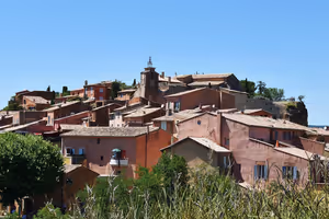 Ocker- und rosafarbene Häuser mit Ziegeldächern, am Hang um einen Glockenturm gestapelt, in Roussillon unter klarem blauem Himmel