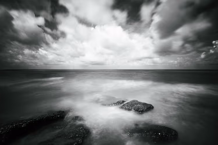 Zwart-witopname met lange sluitertijd van donkere rotsen in nevelig zeewater onder zware, dramatische wolken