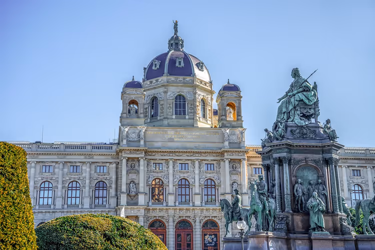 Museumsfassade mit Kuppel und das bronzene Maria-Theresien-Denkmal hinter gestutzten Hecken in Wien bei klarem Himmel