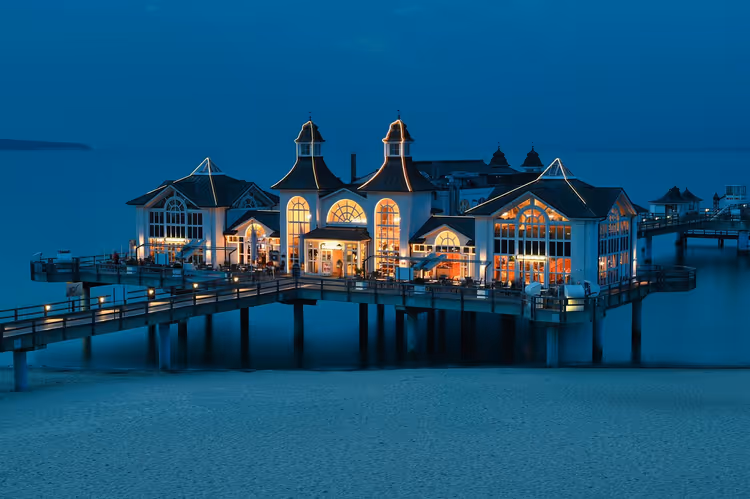 Beleuchteter weißer Seebrücken-Pavillon mit Türmchen auf Stelzen über dem Meer und Sandstrand zur blauen Stunde