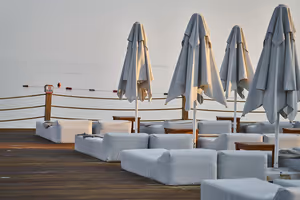 Geschlossene graue Sonnenschirme und weiß gepolsterte Liegen auf einem Holzdeck mit Seilgeländer über ruhigem, dunstigem Meer