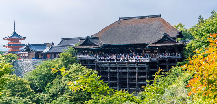 Holztempel mit voller Terrasse auf hohen Stelzen und eine rote Pagode zwischen grünen Bäumen in Japan