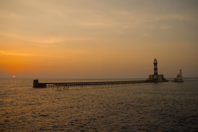 Gestreifter Leuchtturm am Ende eines langen Holzstegs über einem Riff im Roten Meer bei orangefarbenem Sonnenuntergang