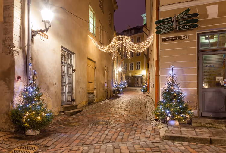 Vicolo acciottolato nella città vecchia di Tallinn di notte con alberi di Natale illuminati, una ghirlanda di luci in alto e una lanterna a muro