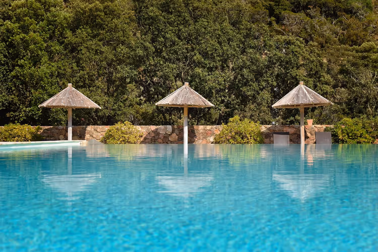 Türkisfarbener Pool mit drei Strohschirmen entlang einer Steinmauer, dahinter dichte grüne Bäume