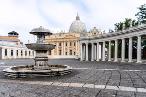 Kleiner mehrstufiger Steinbrunnen auf einem gepflasterten Platz, dahinter eine geschwungene Kolonnade und die Kuppel des Petersdoms