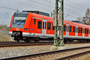 Roter Nahverkehrszug der Deutschen Bahn fährt an einem trüben Tag unter Oberleitungen über die Gleise