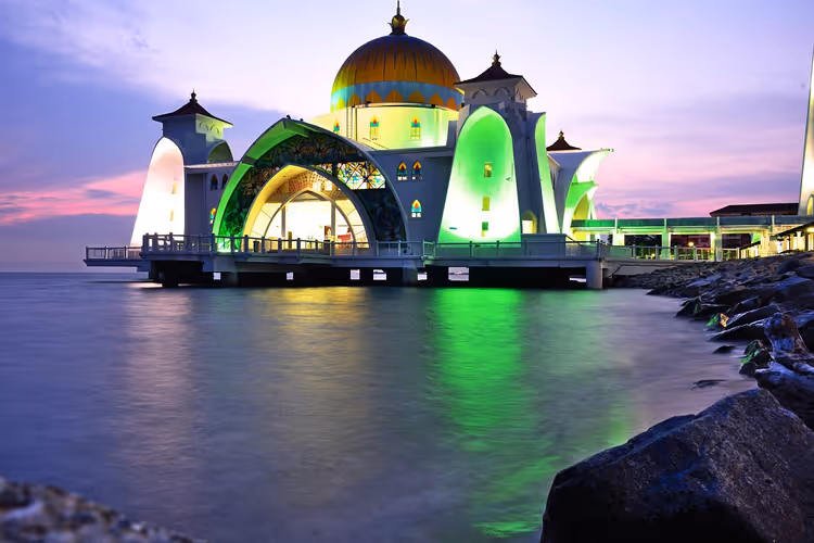 Moschee auf Stelzen über dem Meer, in der Dämmerung grün beleuchtet, mit goldener Kuppel und felsigem Ufer, Malaysia