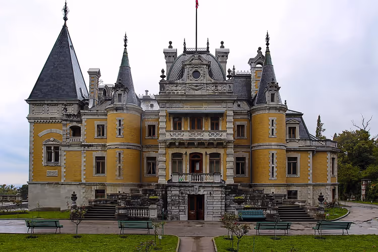 Gelber Palast im Stil eines Châteaus mit spitzen Schiefertürmchen und Gartenbänken unter bedecktem Himmel bei Jalta