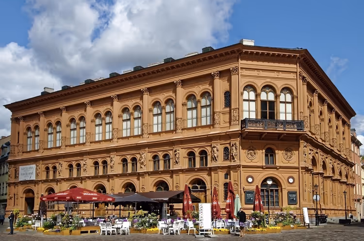 Reich verziertes terrakottafarbenes Neorenaissance-Gebäude in Riga mit Bogenfenstern und Cafétischen auf dem Platz