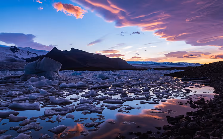 Eisbrocken treiben in der Gletscherlagune Jökulsárlón, dunkle Berge und rosa Abendwolken spiegeln sich im Wasser