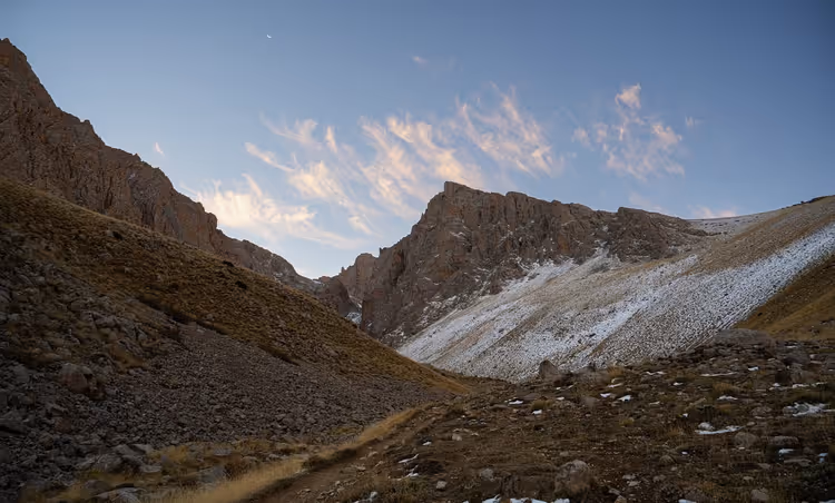 Felsiges Bergtal in der Dämmerung mit Schneefeldern an den Hängen und rosa Wolken über einem gezackten Grat