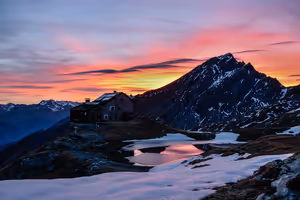 Berghütte an einem kleinen Teich mit Schneerändern unter rosa-orangem Abendhimmel, dahinter ein felsiger Gipfel