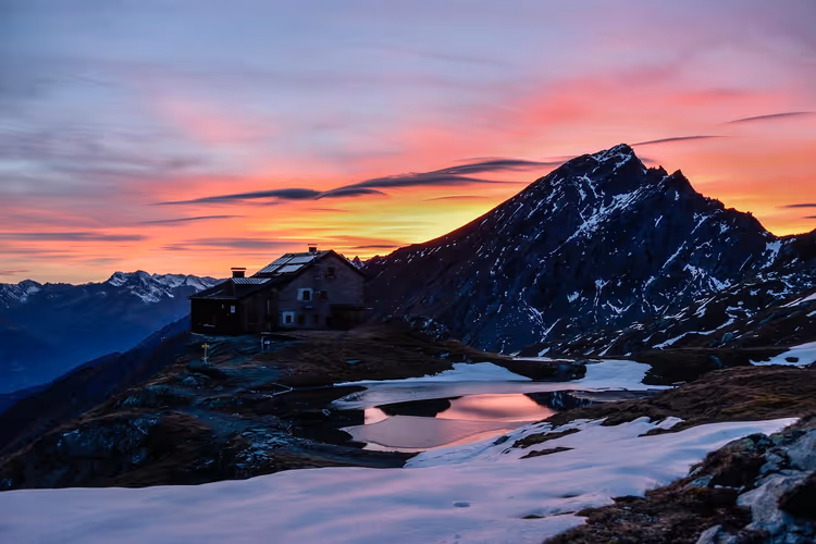 Berghütte an einem kleinen Teich mit Schneerändern unter rosa-orangem Abendhimmel, dahinter ein felsiger Gipfel