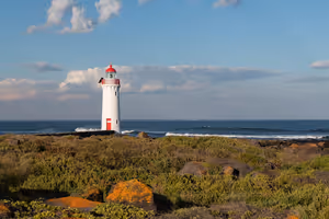 Weißer Leuchtturm mit roter Spitze an einer felsigen, grasbewachsenen Küste in Australien, dahinter brechende Ozeanwellen