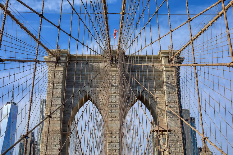 Steinturm der Brooklyn Bridge mit Spitzbögen, gesehen durch ein Netz aus Tragseilen vor blauem Himmel