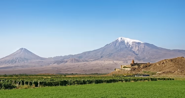 Kloster Chor Virap auf einem niedrigen Hügel über grünen Weinbergen, dahinter die schneebedeckten Gipfel des Ararat