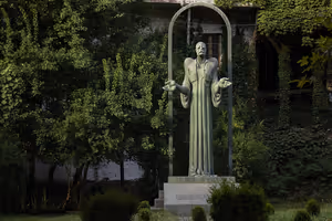 Hohe grüne Bronzestatue eines Mannes im Gewand mit ausgestreckten Händen unter einem Metallbogen in einem efeubewachsenen Garten