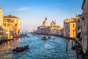 Breiter Kanal mit pastellfarbenen Palazzi und einer Kuppelkirche, Boote und ein Wasserbus auf dem Wasser im warmen Licht