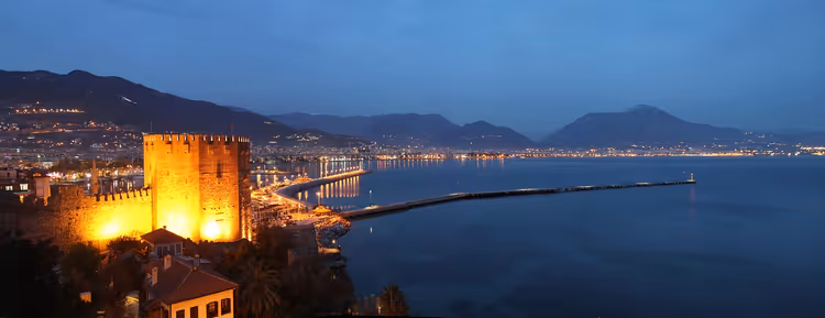 Beleuchteter Roter Turm über dem Hafen von Alanya in der Dämmerung, mit langem Pier, Stadtlichtern und Bergen jenseits der Bucht