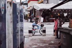 Frau fährt in einer Seitenstraße in Singapur auf dem Fahrrad an gestreiften Pollern vorbei, zwei Menschen schauen vom Gehweg aus zu