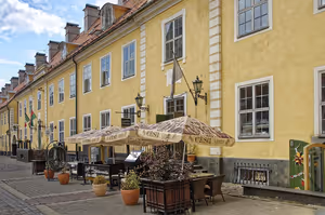 Riga'nın eski şehrinde kiremit çatılı uzun sarı tarihi bina, önünde şemsiyeli ve saksı bitkili bir kafe terası