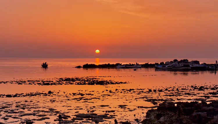Orangefarbener Sonnenuntergang über ruhigem Meer auf Djerba, ein kleines Boot und eine felsige Landzunge als Silhouetten vor dem leuchtenden Wasser