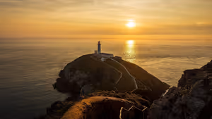 Phare de South Stack sur un îlot rocheux au large d'Anglesey au couchant, avec un sentier sinueux et une lumière dorée sur la mer