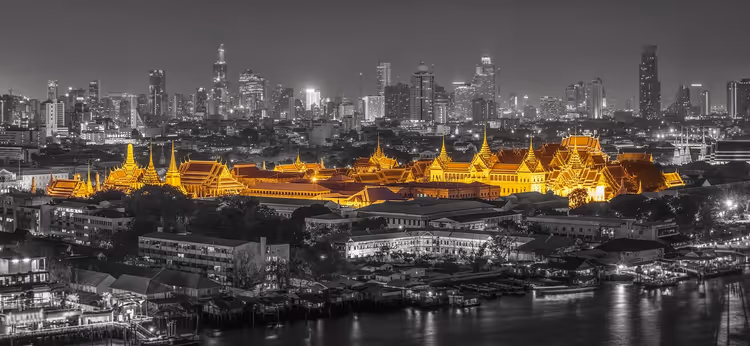 Großer Palast in Bangkok, nachts golden leuchtend vor einer schwarz-weißen Stadtsilhouette, davor der Fluss