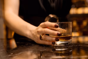 Hand mit dunkelroten Nägeln hält ein geriffeltes Glas Whiskey auf einem polierten Marmortresen in warmem, gedämpftem Licht