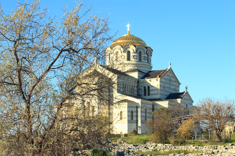Gestreifte Steinkathedrale mit vergoldeter Kuppel über antiken Ruinen und kahlen Frühlingsbäumen in Sewastopol