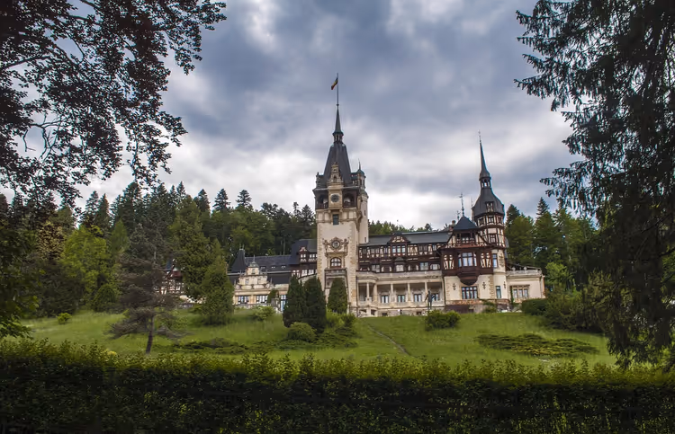 Schloss Peleș in Sinaia mit spitzen Türmen und Fachwerkfassaden an einem grünen Hang vor Tannenwald und Wolken
