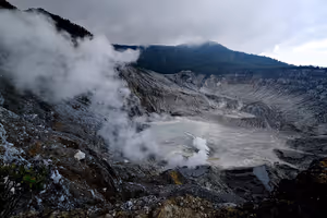 Dampf steigt aus einem grauen Vulkankrater mit einem blassen Schwefelsee am Grund unter schweren Wolken in Indonesien