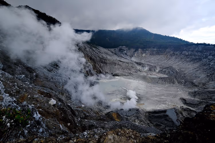 Dampf steigt aus einem grauen Vulkankrater mit einem blassen Schwefelsee am Grund unter schweren Wolken in Indonesien