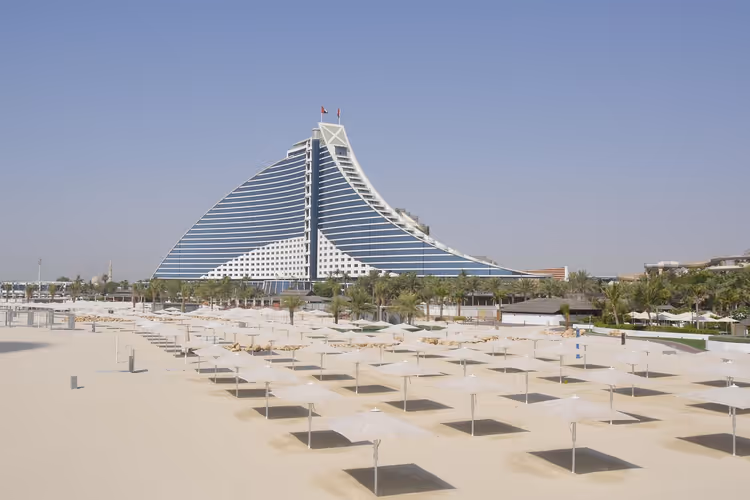 Wellenförmiger Hotelturm hinter einem breiten Sandstrand mit Reihen weißer Sonnenschirme in Dubai