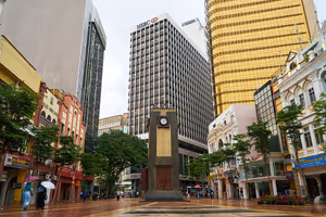 Uhrturm auf einem regennassen Fußgängerplatz in Malaysia, umrahmt von kolonialen Ladenhäusern und Bürotürmen