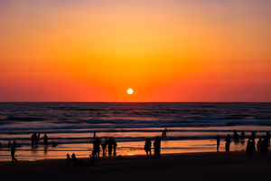 Silhouetten von Menschen an einem Strand in Goa bei Sonnenuntergang, die Sonne steht tief über dem Meer unter orangefarbenem Himmel